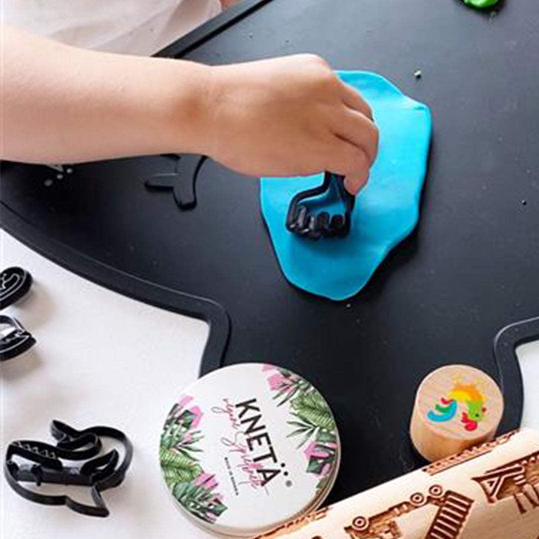 Een kinderhand drukt een zwarte Knetä giraf uitsteekvorm in blauwe klei op een zwarte speelmat.