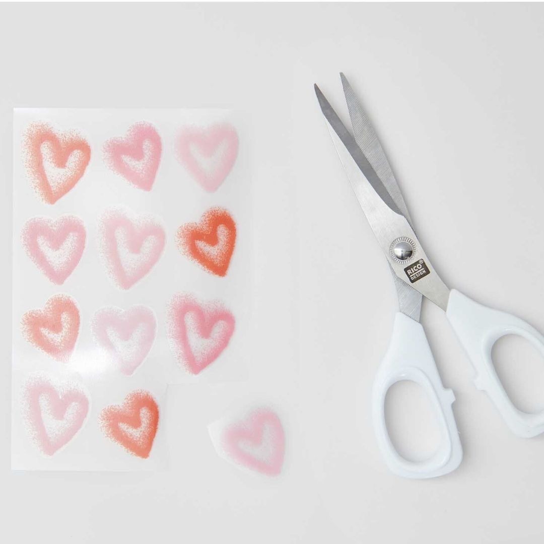 Een vel Rico Design transferfolie met roze en rode spray harten en een schaar ernaast.