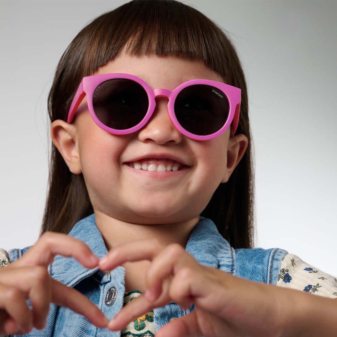 Een lachend kind draagt de roze Komono kinderzonnebril en maakt een hartje met haar handen.