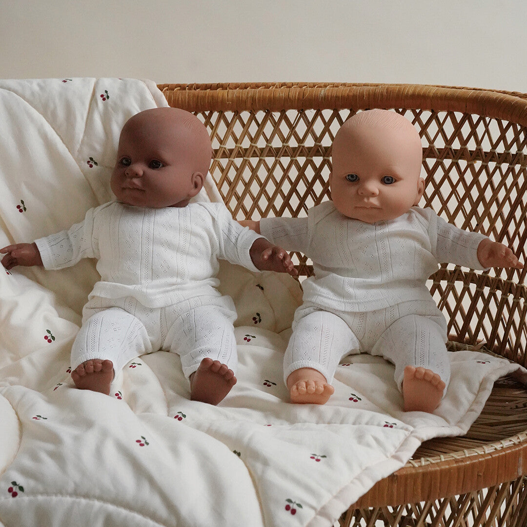 Twee Konges Sløjd babypoppen, één met lichte en één met donkere huid, zittend op een stoel in witte gebreide pakjes.