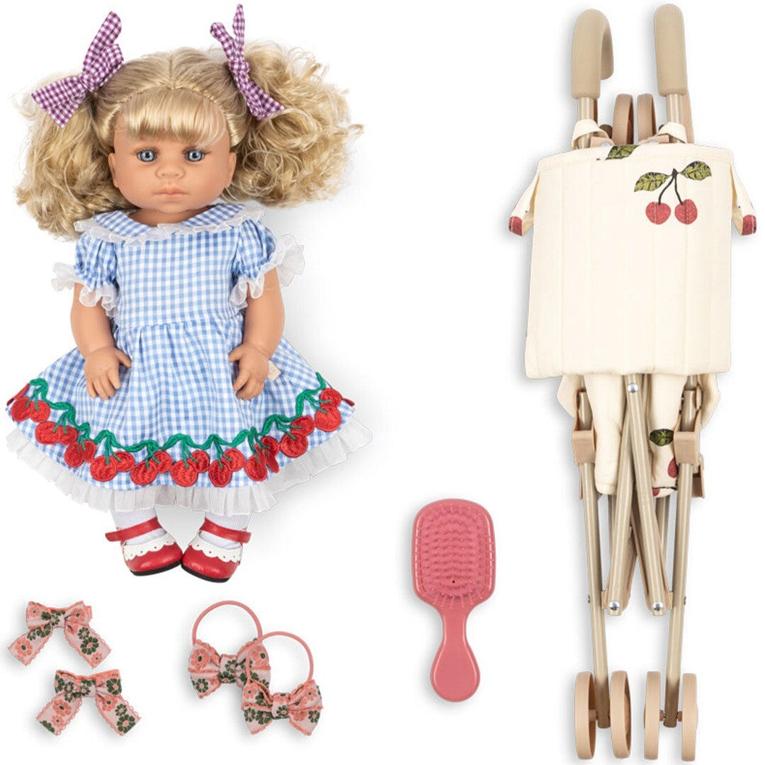 Konges Sløjd babypop Delphine met blauw geruite jurk, een opgevouwen buggy, roze borstel en haarstrikjes, liggend op een witte achtergrond.