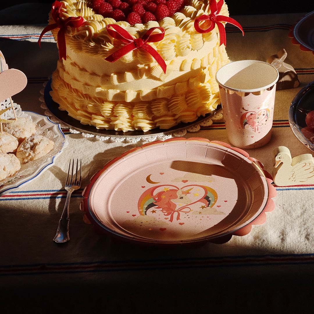 Een feesttafel met een taart, koekjes, en een roze papieren beker en bord met een eenhoorn- en regenboogprint.