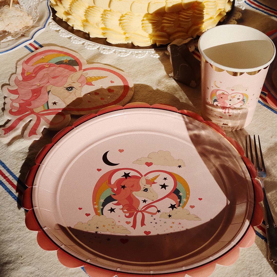 Een close-up van een feesttafel met een roze papieren bord, servet en beker met een eenhoorn- en regenboogdesign.
