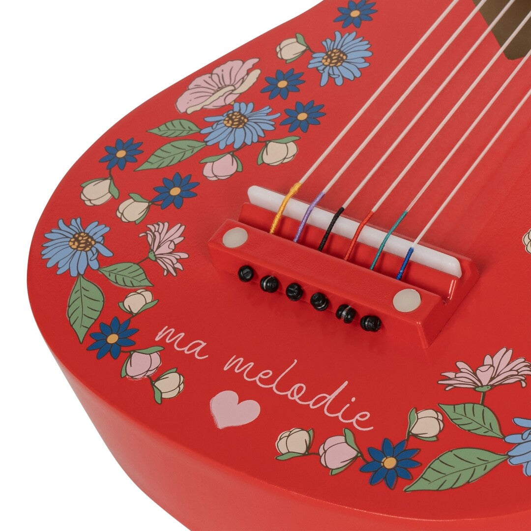 Detail van de rode speelgoedgitaar met de brug, gekleurde snaren, bloemen en de tekst 'ma melodie'.