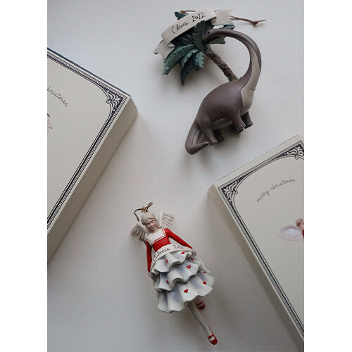 Twee Konges Sløjd kersthangers, een dinosaurus met palmboom en een engel, liggen naast twee kerstdozen.