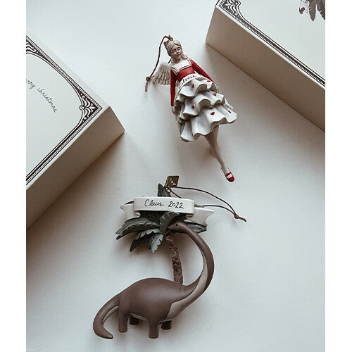Twee Konges Sløjd kersthangers, een dinosaurus met palmboom en een engel, liggen naast twee kerstdozen.