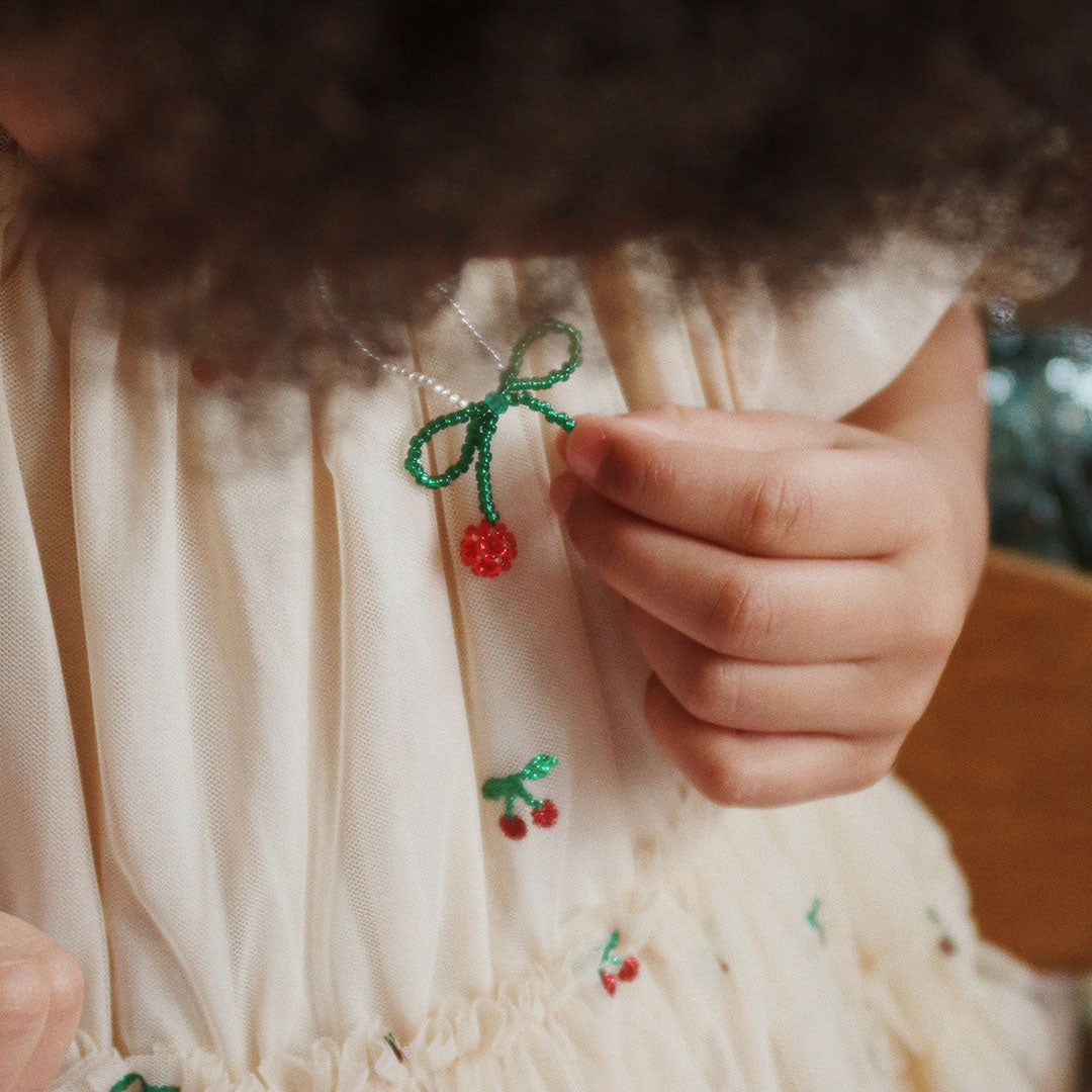 Een kinderhand houdt de Konges Sløjd kersenketting vast, gedragen over een crèmekleurige jurk.