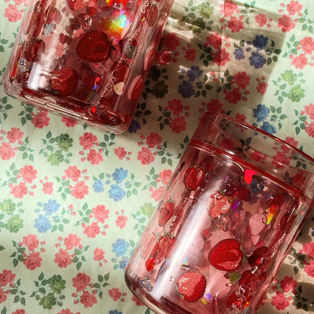Twee Konges Sløjd kinderbekers met aardbeien en glitter op een bloemrijk tafelkleed.