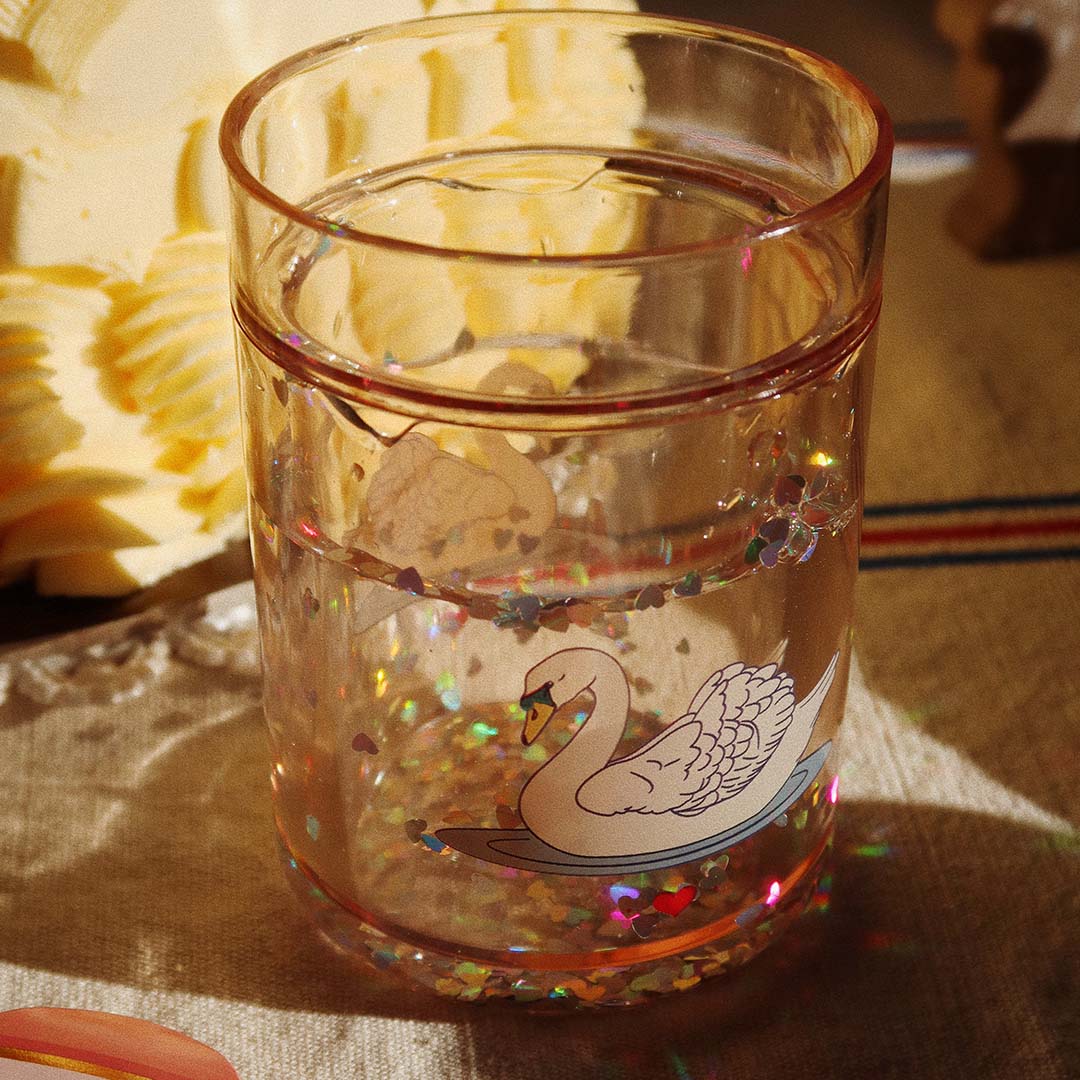 Een Konges Sløjd kinderbeker met zwaan en glitter op een tafel met warme belichting.