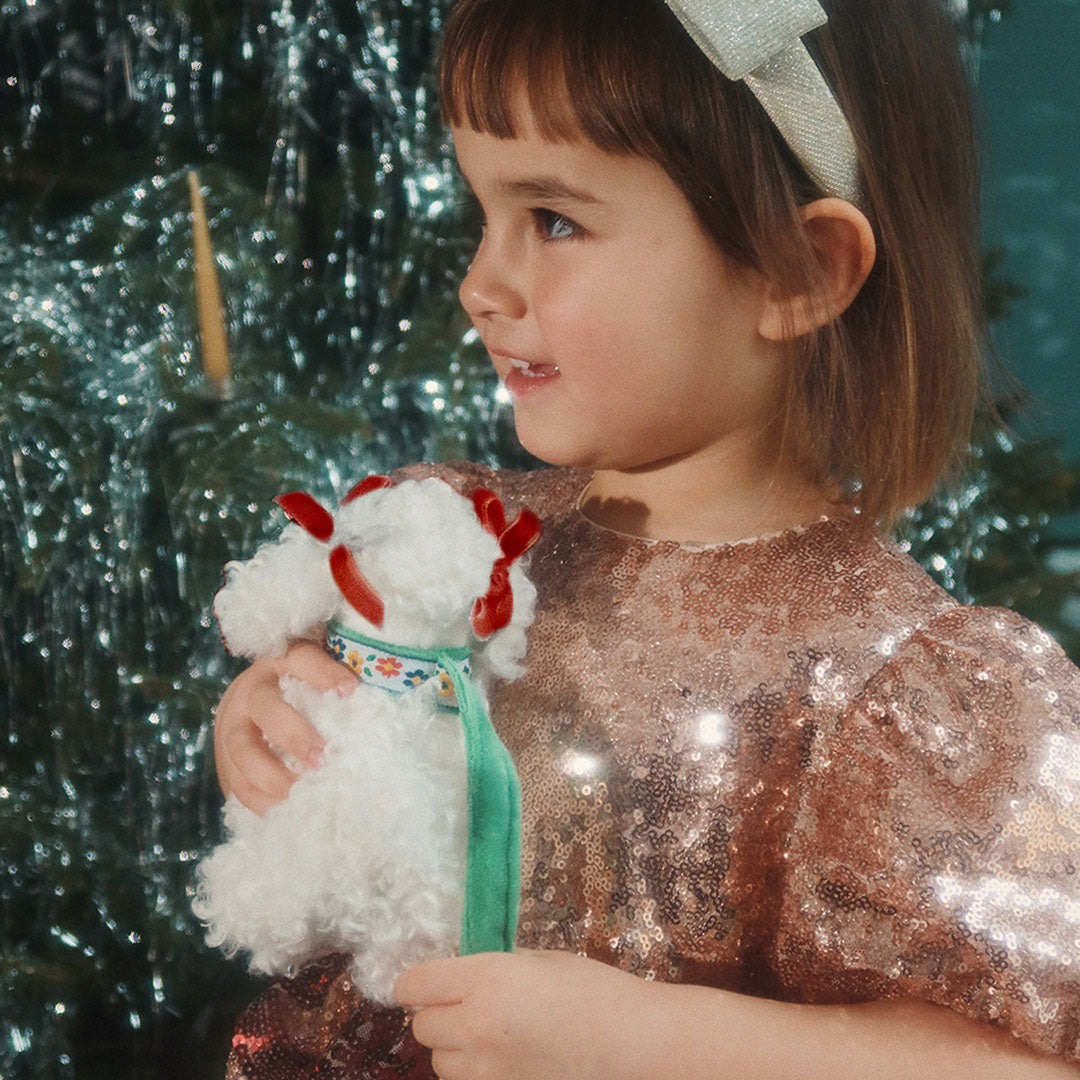 Een kind houdt de Konges Sløjd witte pluchen poedel knuffelpuppy vast voor een glimmende kerstboom.
