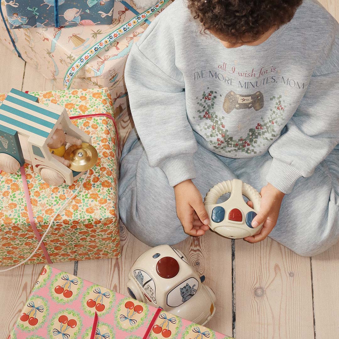 Een kind speelt met de op afstand bestuurbare auto met kattenprint en afstandsbediening tussen cadeaus op een houten vloer.