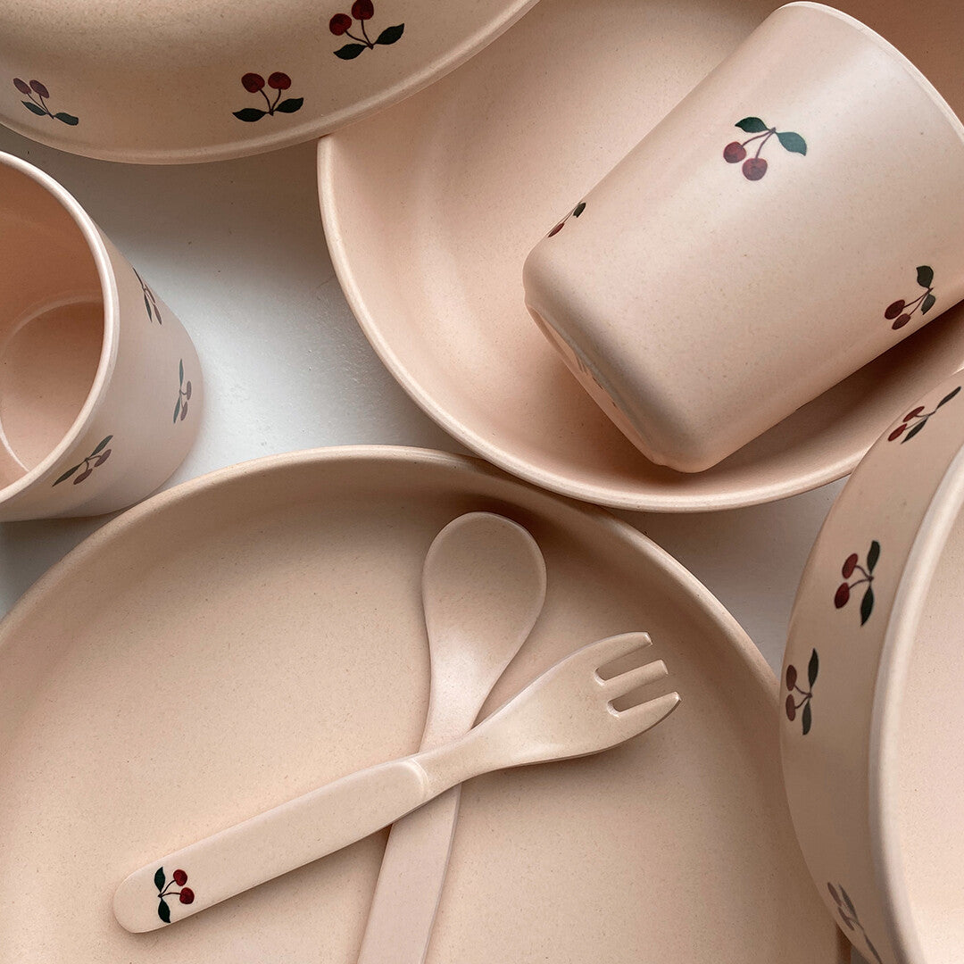 Detailopname van het beige kinderservies met kersjesprint, met een bord, kom, beker, vork en lepel.