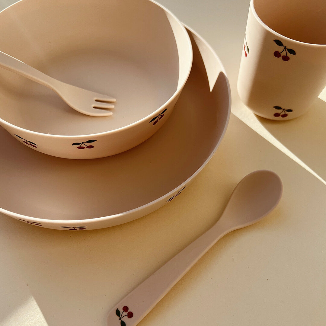 Beige kinderservies met kersjesprint, met een bord, kom, beker en lepel, in natuurlijk licht.