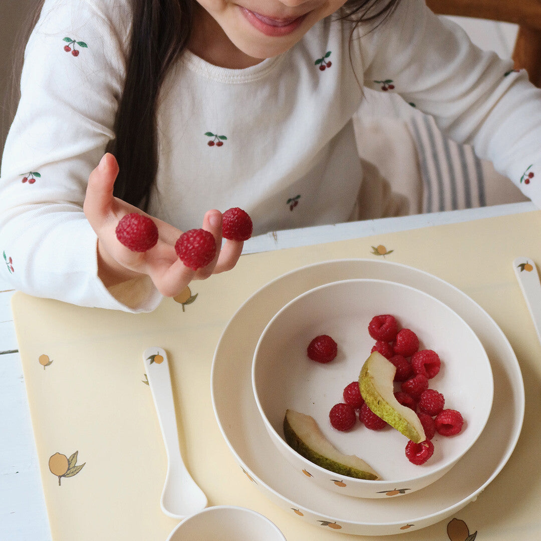 Een kind dat frambozen vasthoudt boven een kom met fruit op het Konges Sløjd beige kinderservies met citroenprint.