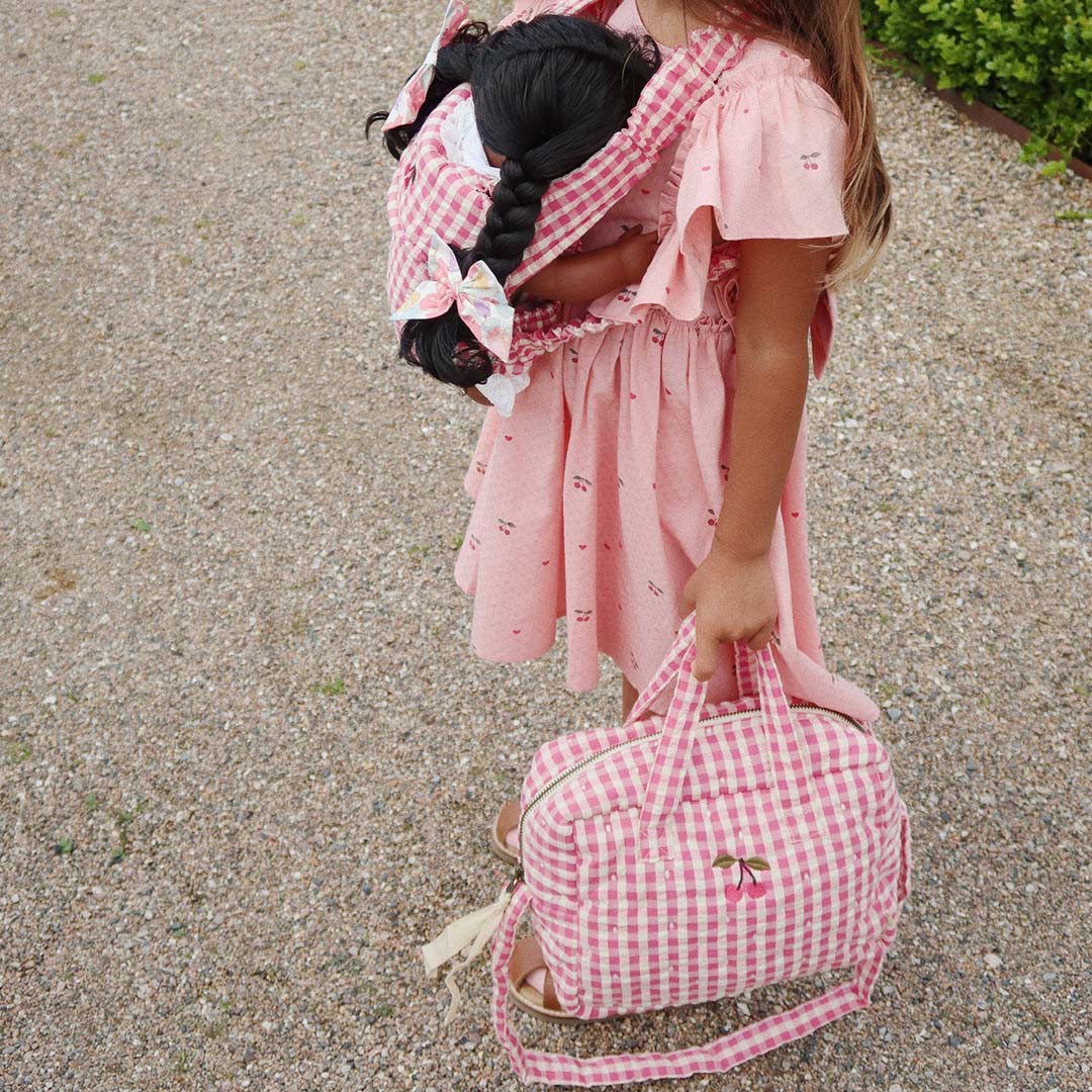 Een kind draagt de Konges Sløjd poppenluiertas met roze-wit ruitpatroon en een pop in een bijpassende draagzak.