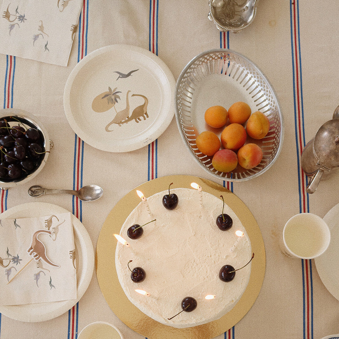 Een feestelijk gedekte tafel met Konges Sløjd papieren servetten en bordjes met dinosauruspatroon, een taart en fruit.