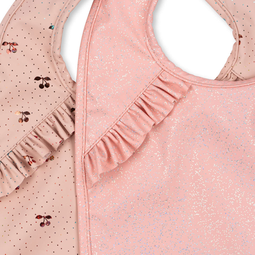 Detailopname van de roze babyslabbetjes met ruches, één met kersenpatroon en één met glitters.