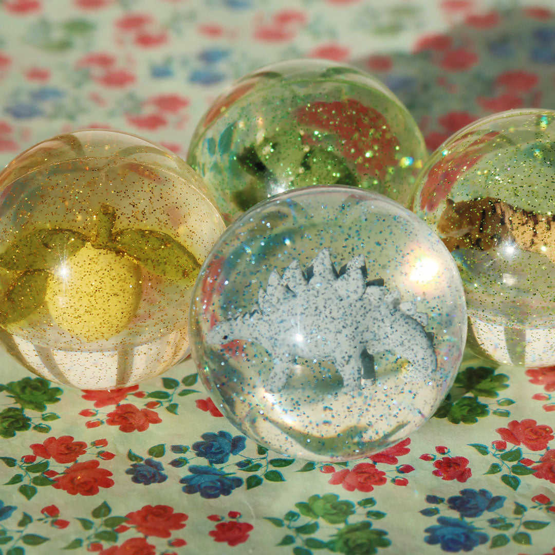 Vier dinosaurus stuiterballen met glitter op een bloemige ondergrond.