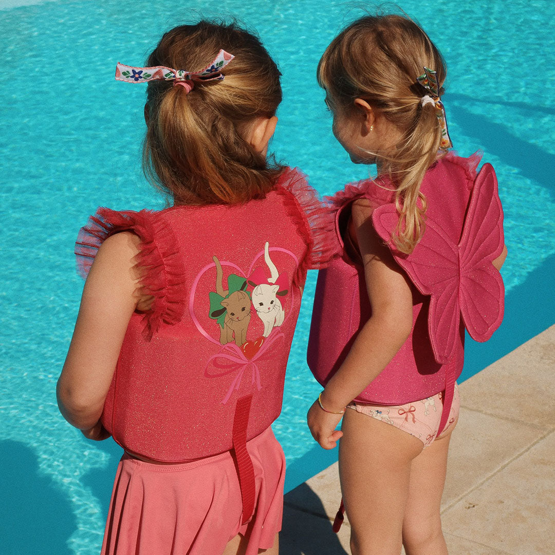 Twee kinderen met zwemvesten bij een zwembad, waarvan één met een rood zwemvest met kattenprint en de ander met een roze zwemvest met vlindervleugels.