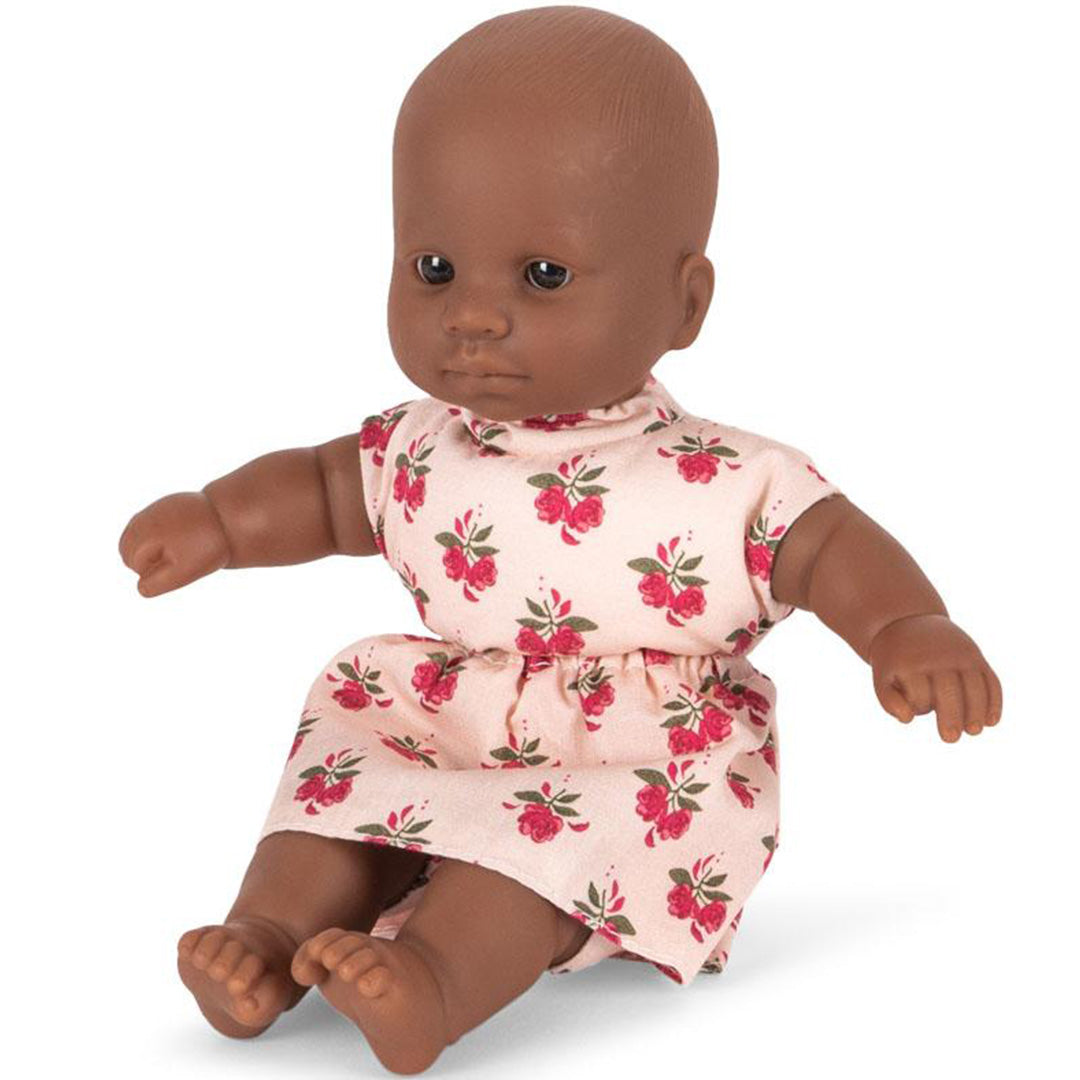 Konges Sløjd babypop met donkere huidskleur, zittend in een roze jurkje met gebloemd patroon.