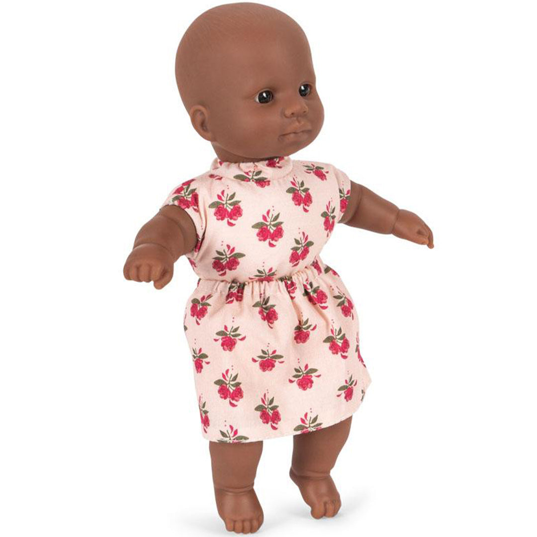 Konges Sløjd babypop met donkere huidskleur, staand en schuin gefotografeerd in een roze jurkje met gebloemd patroon.