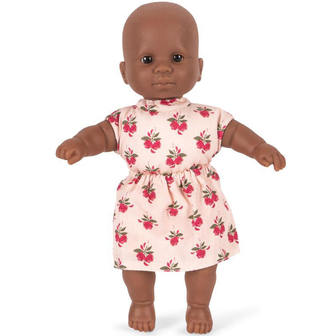 Konges Sløjd babypop met donkere huidskleur, staand in een roze jurkje met gebloemd patroon.