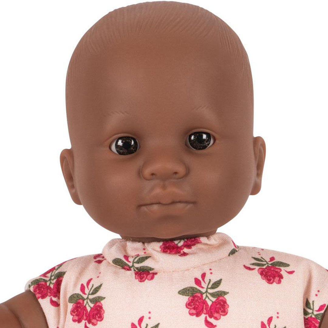 Close-up van het gezicht van de Konges Sløjd babypop met donkere huidskleur en een roze jurkje met gebloemd patroon.