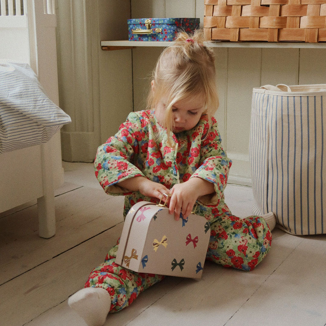 Een kind in bloemenpyjama opent de beige Konges Sløjd kinderkoffer met strikjes op de vloer.