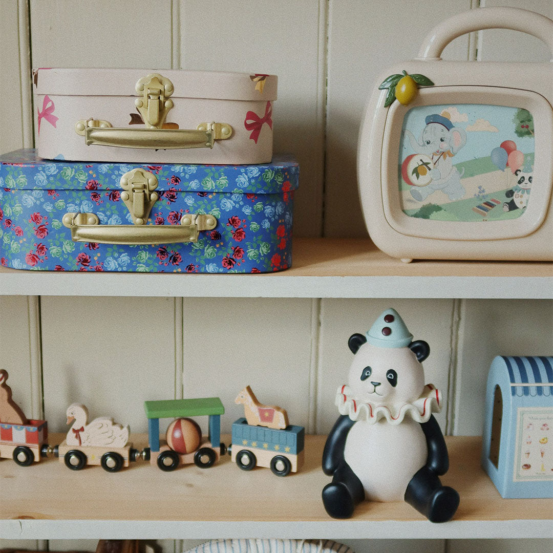 De twee Konges Sløjd kinderkoffers, beige met strikjes en blauw met bloemen, staan op een houten plank tussen ander speelgoed.