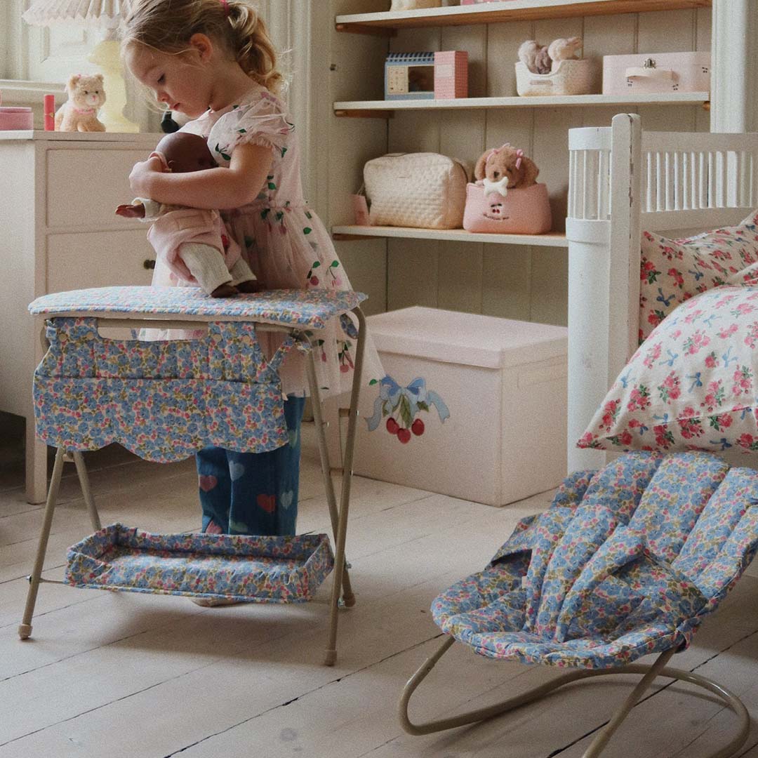 Een kind houdt een pop vast naast de Konges Sløjd poppenverschoontafel met gebloemd patroon in een kinderkamer.