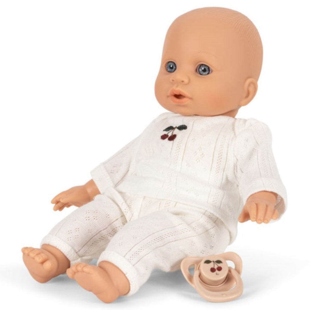 Konges Sløjd babypop met blauwe ogen in een wit gebreid pakje, zittend naast een beige speen.