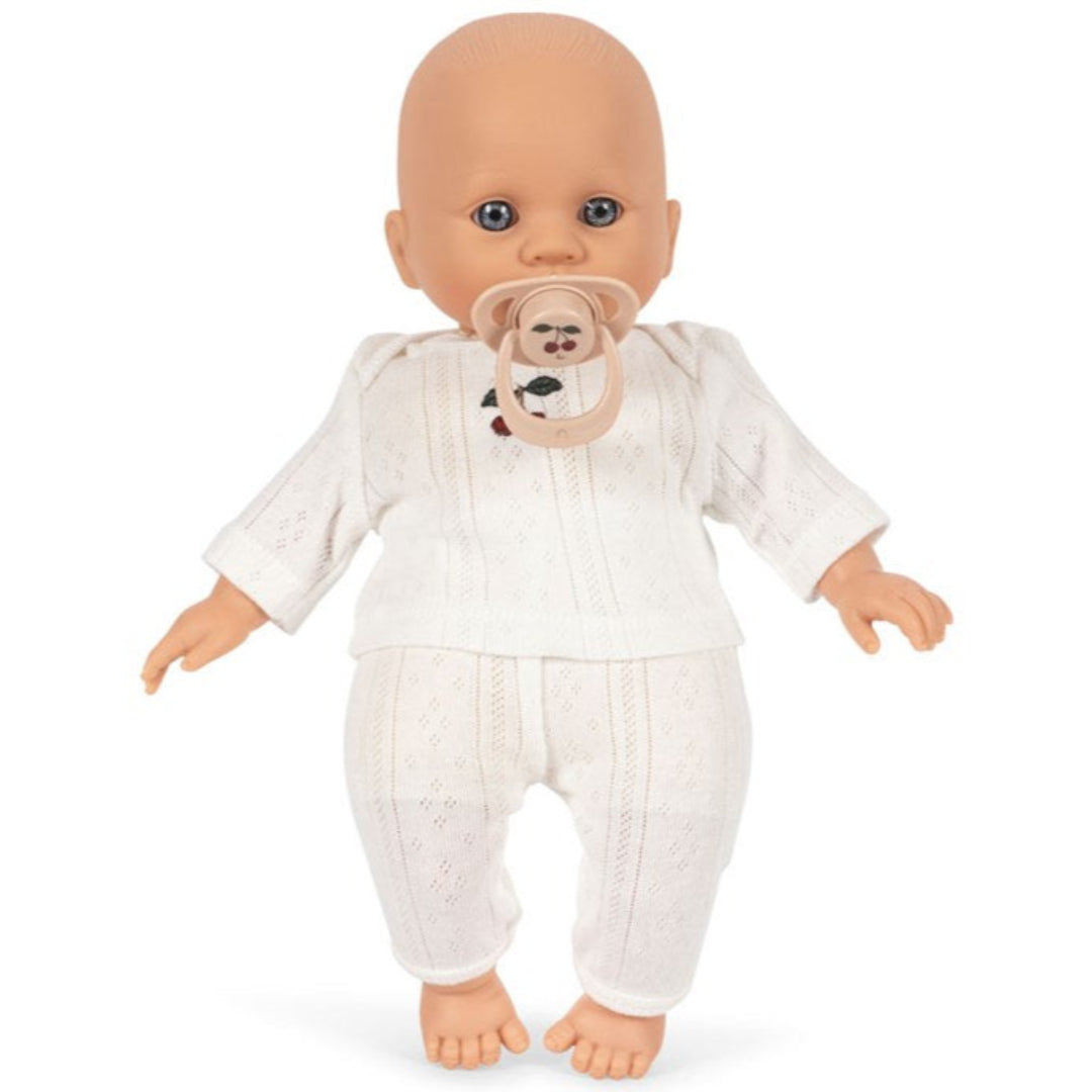 Konges Sløjd babypop met blauwe ogen in een wit gebreid pakje, staand met een beige speen in de mond.
