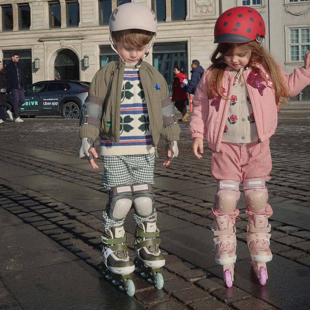 Twee kinderen met Konges Sløjd kinderskeelers en helmen, staand op een kasseienweg.