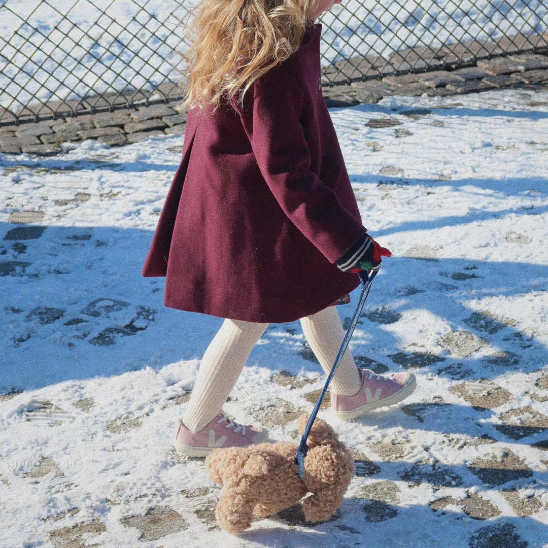 Een kind loopt in de sneeuw en trekt een bruine knuffelpuppy aan een blauw geruite riem voort.