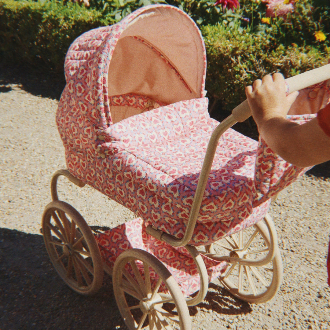 Een kind duwt een Konges Sløjd poppenwagen met roze hartenpatroon over een pad.