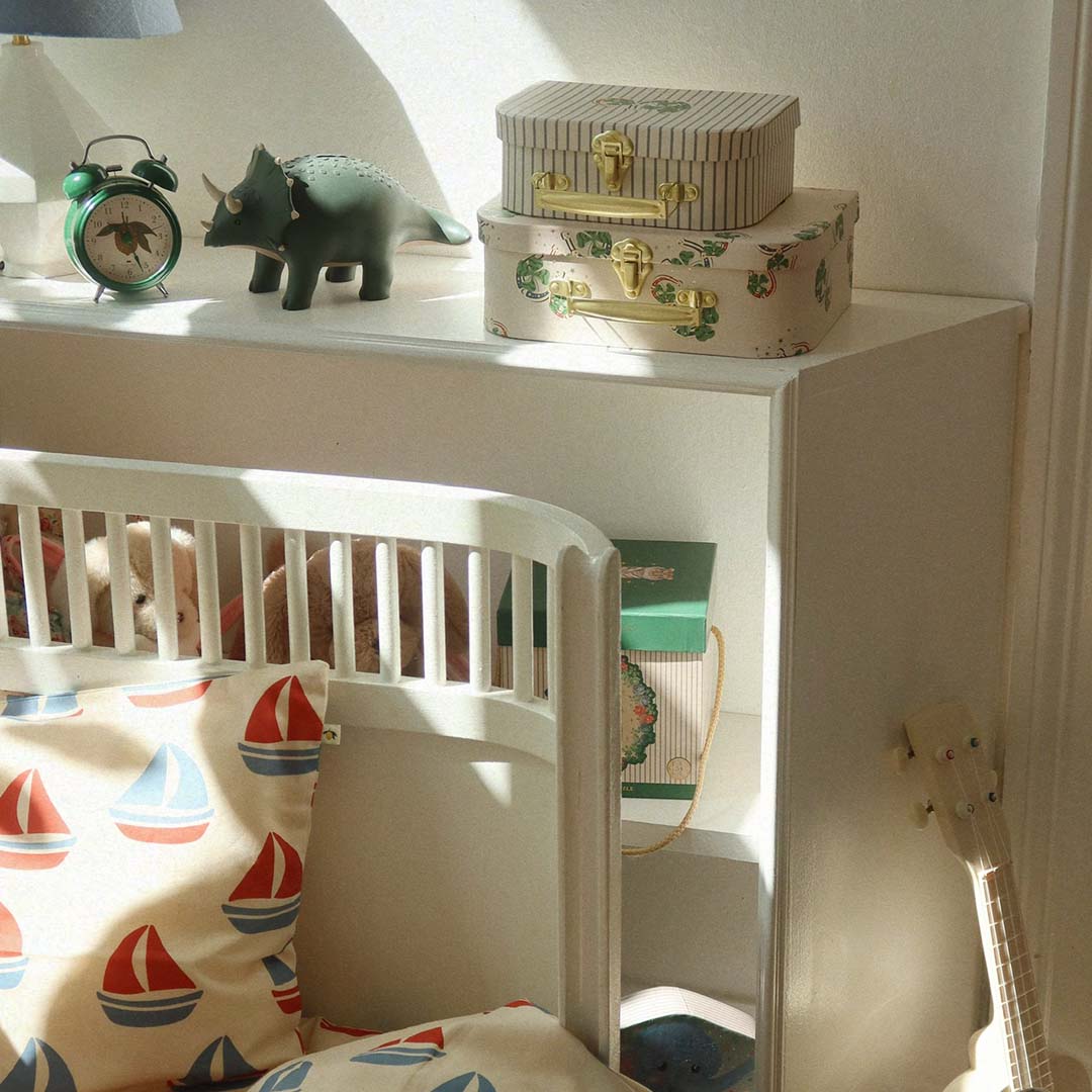 De groene wekker staat op een plank in een kinderkamer, naast een dinosaurus en opbergdozen.