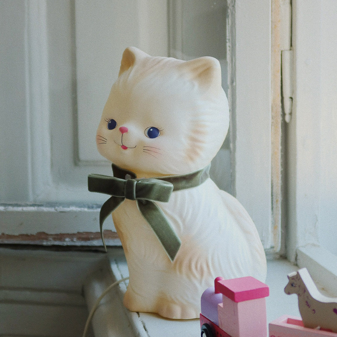 Konges Sløjd kinderlamp in de vorm van een beige kat met groene strik, staand op een vensterbank naast houten speelgoed.