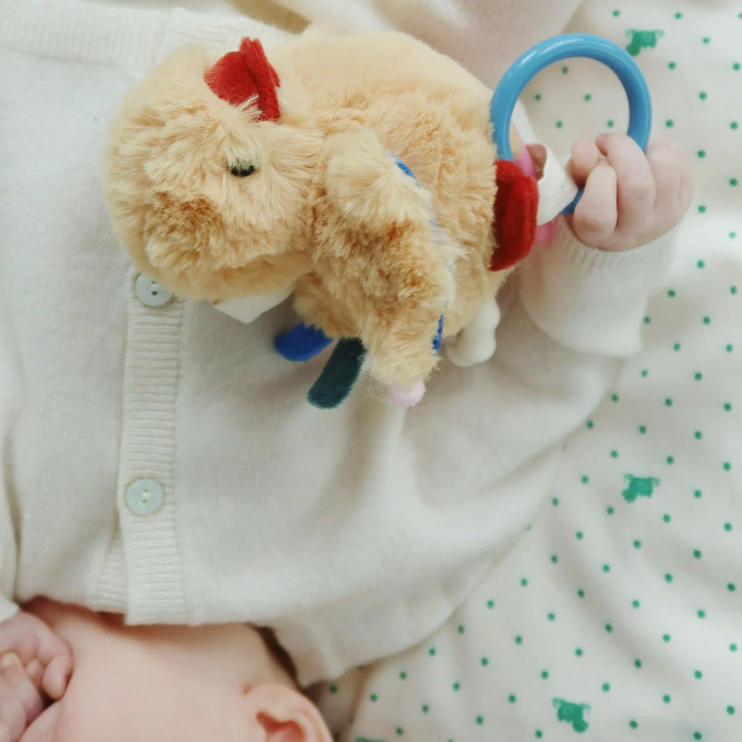 Een babyhandje houdt de Konges Sløjd activiteitenknuffel kuiken vast, liggend op een witte cardigan en een groen gestippelde deken.