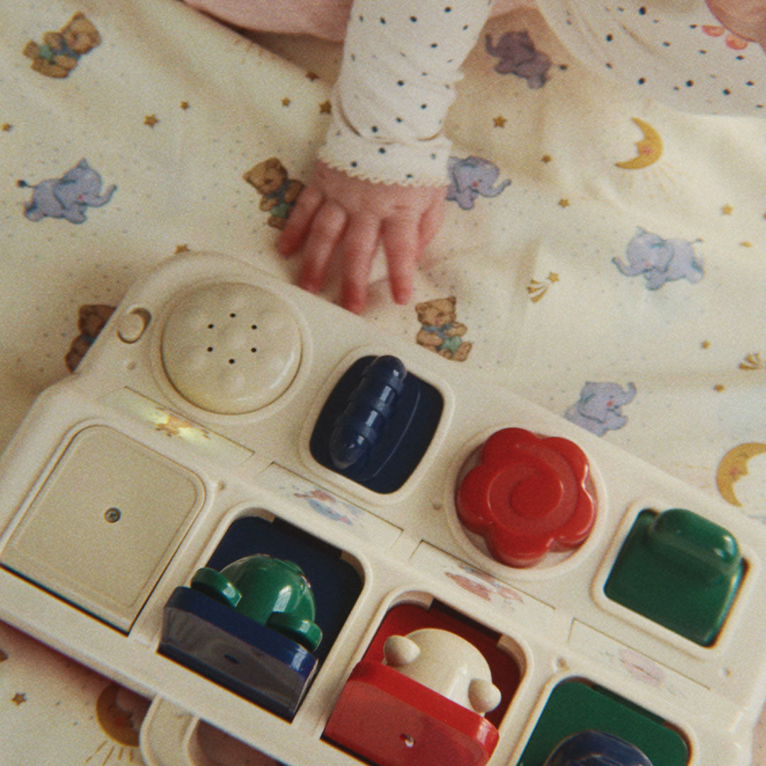 Een babyhandje speelt met het Konges Sløjd activiteitenpaneel met geluid, dat op een deken met dierenpatroon ligt.