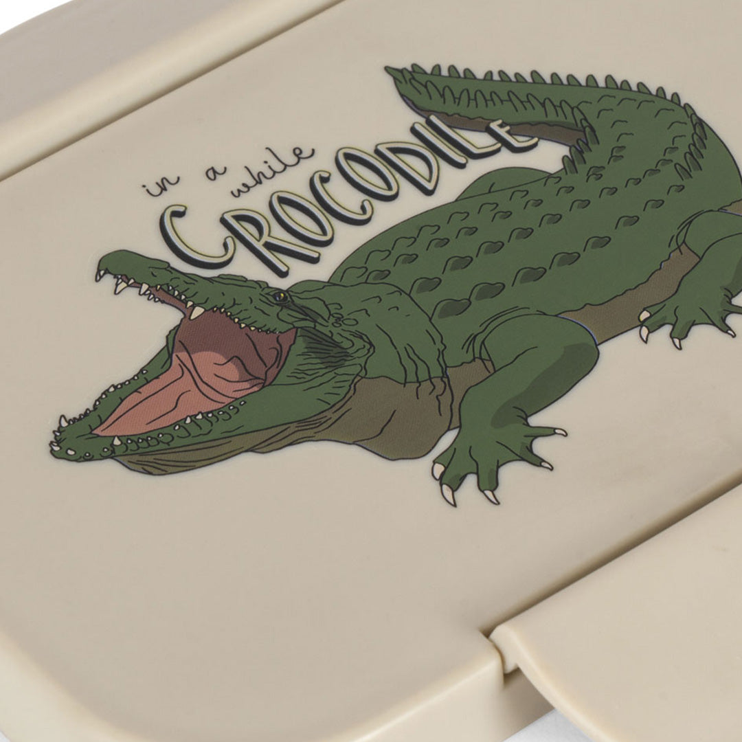 Detail van de deksel van de broodtrommel met een illustratie van een groene krokodil en de tekst 'in a while CROCODILE'.
