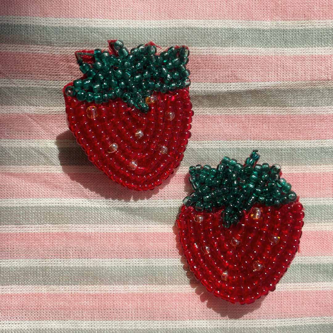 Twee Konges Sløjd haarclips in de vorm van aardbeien, gemaakt van rode en groene kraaltjes, liggend op een gestreepte stof.