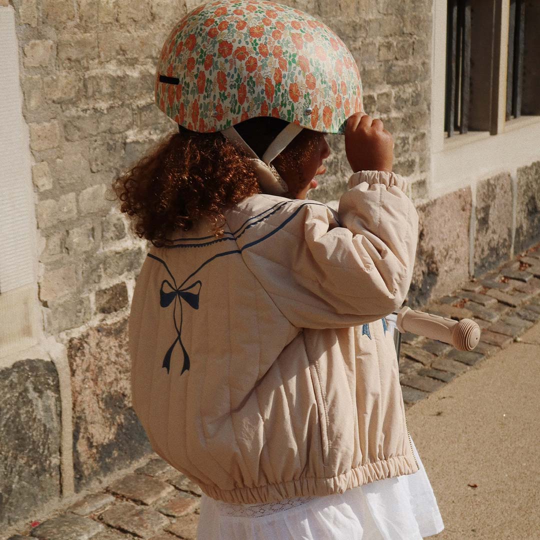 Een kind met krullend haar draagt de Konges Sløjd kinderhelm met gebloemd patroon en raakt de helm aan.