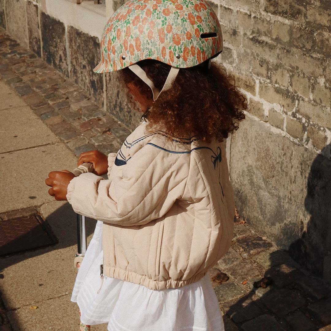 Een kind draagt de Konges Sløjd kinderhelm met gebloemd patroon en rijdt op een step.