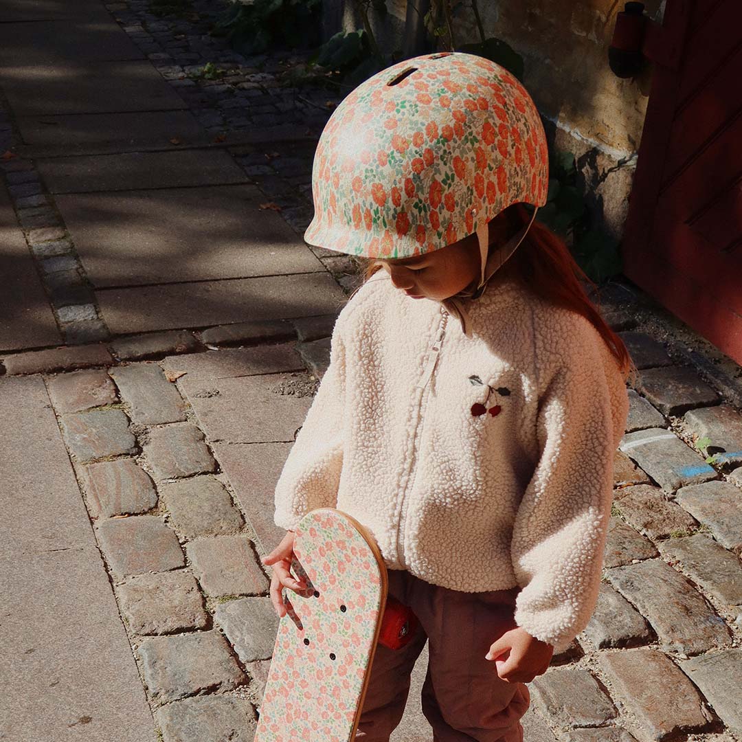 Een kind draagt de Konges Sløjd kinderhelm met gebloemd patroon en houdt een skateboard vast.