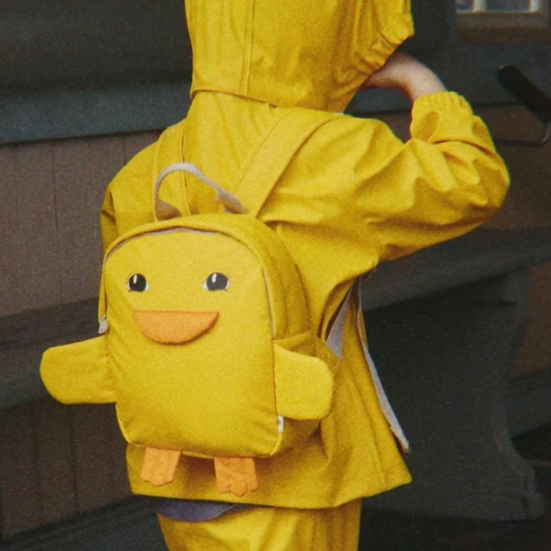 Een kind met gele regenjas draagt de gele eenden peuterrugzak op de rug.