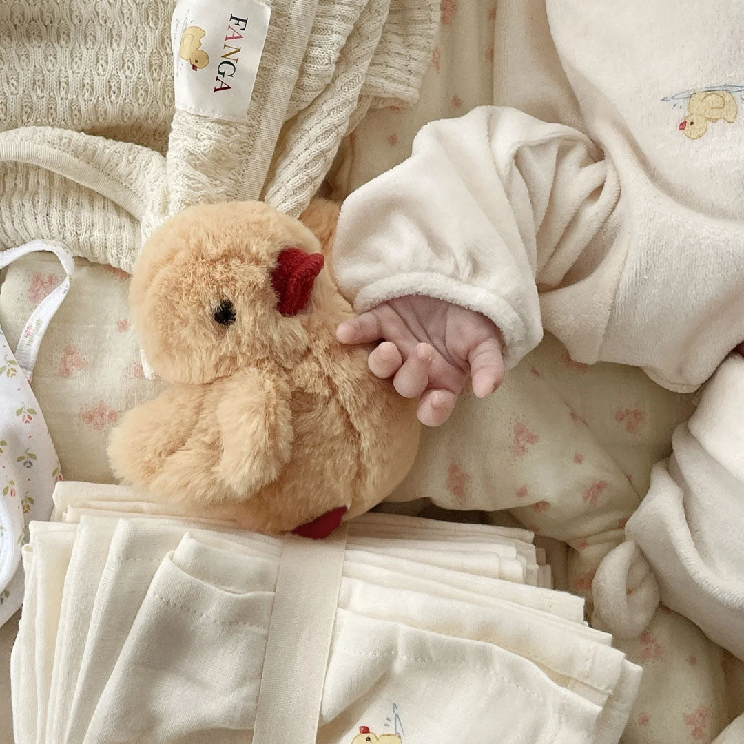 Een babyhandje raakt de Konges Sløjd rammelaar knuffelkuiken aan, liggend tussen zachte babykleding.