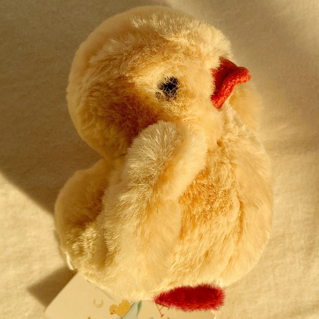 Zijaanzicht van de Konges Sløjd rammelaar knuffelkuiken met een productlabel eronder.