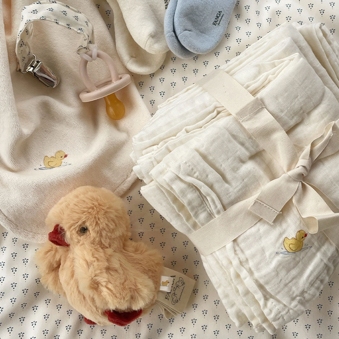 De Konges Sløjd rammelaar knuffelkuiken ligt tussen diverse babyartikelen zoals een speen, sokken en hydrofiele doeken.