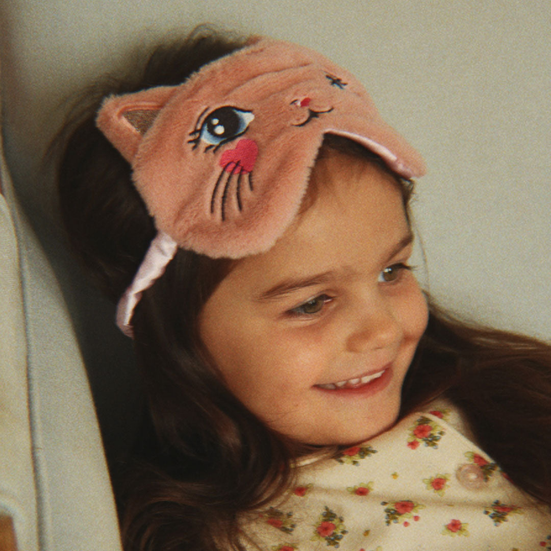 Een lachend meisje met lang haar draagt het roze katten slaapmasker op haar voorhoofd.
