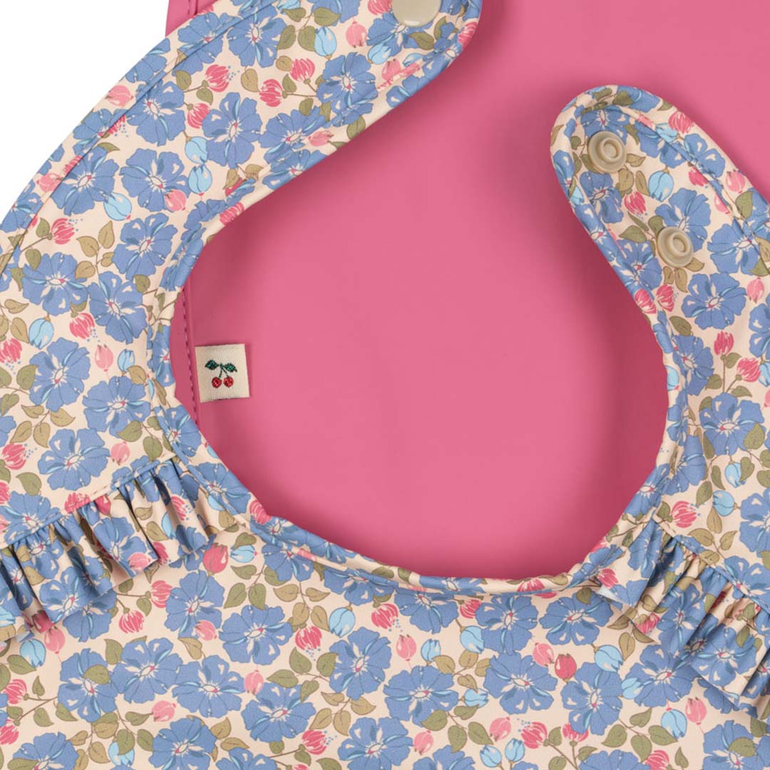 Detail van een Konges Sløjd baby slabbetje met blauw bloemenpatroon en ruches, met een roze slabbetje eronder.